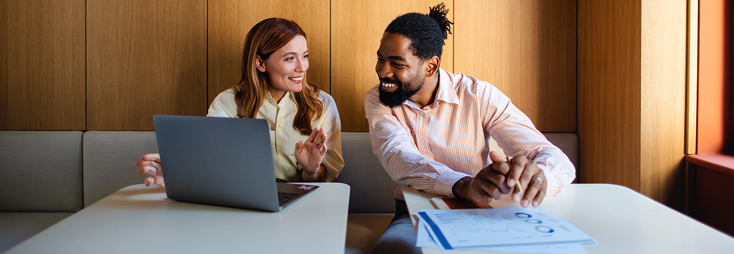 A man and woman are at a table, using a laptop, appearing to collaborate or engage in a discussion.